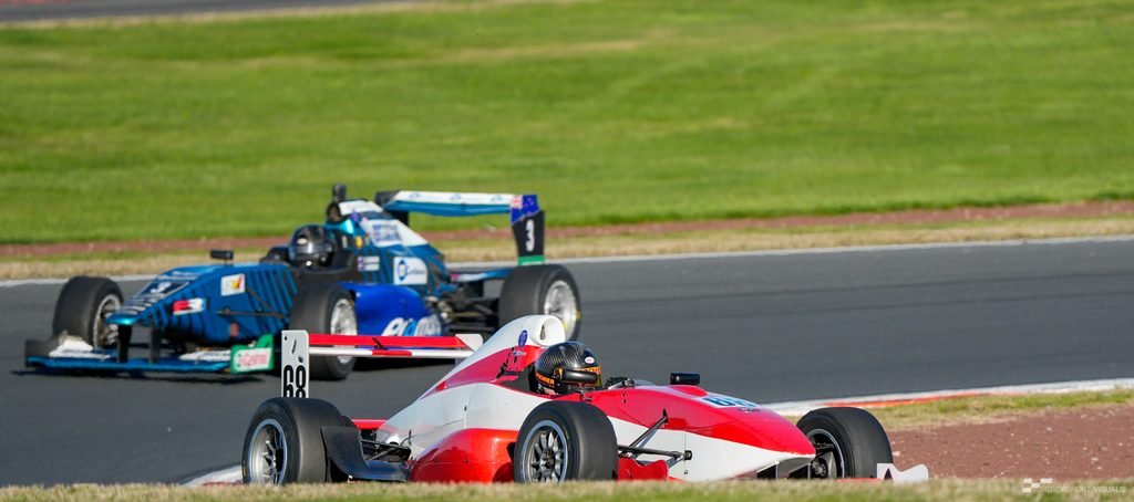 A low-angle action shot captures a race car, likely a Formula racing machine, speeding on a track. The primary subject is the red and white car in the foreground, with another blue race car following behind on a parallel section of the circuit. The background consists of lush green grass and asphalt track surfaces under bright daylight.