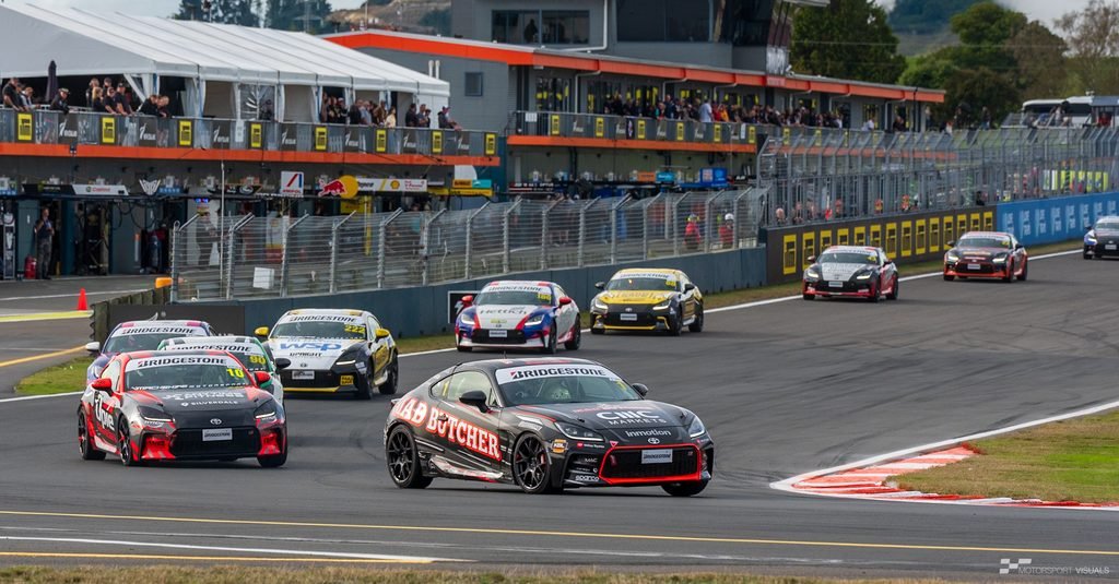 A race scene featuring several high-performance race cars on a track, with grandstands and pit structures in the background. The main focus is a black race car prominently displayed on the asphalt.