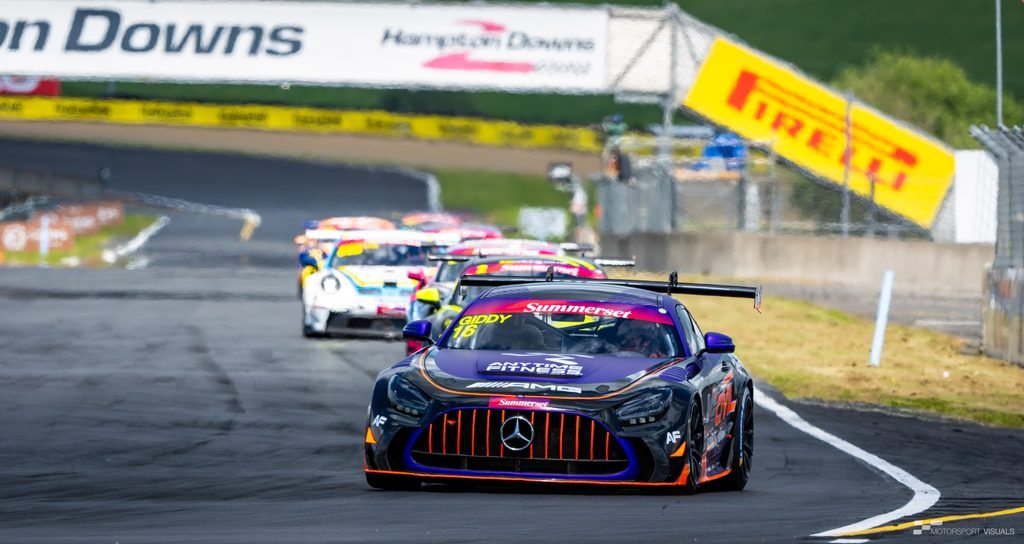 A Mercedes-AMG GT3 race car, painted in black and blue with orange accents, speeds around a racetrack corner during a motorsports event. The background features grandstands and signage for 'Hampton Downs' and 'PIRELLI', indicating a professional racing environment.