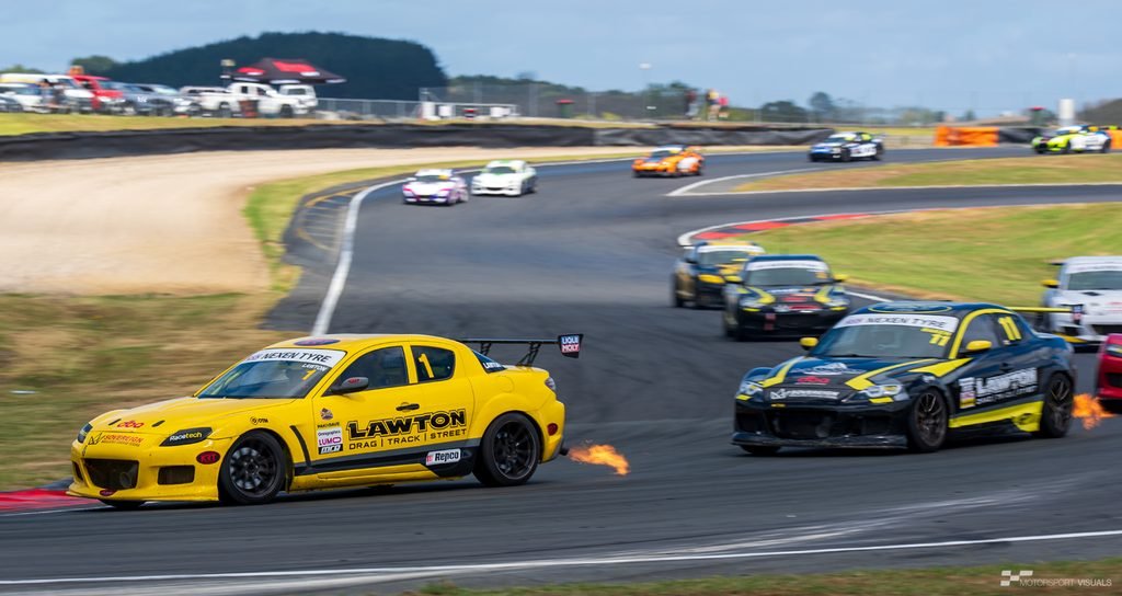 Two yellow and black race cars, likely drag racers or circuit racers, are speeding down a paved track on a sunny day. The cars are kicking up tire smoke and appear to be competing or running side-by-side on a large, winding circuit surrounded by grassy embankments and safety barriers. In the background, there is a wide expanse of open land with distant trees and structures under a bright blue sky.