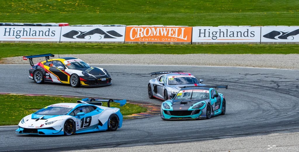 A high-angle action shot captures three race cars navigating a winding asphalt track, with a backdrop featuring signage for 'Cromwell Highlands' and lush green fields.