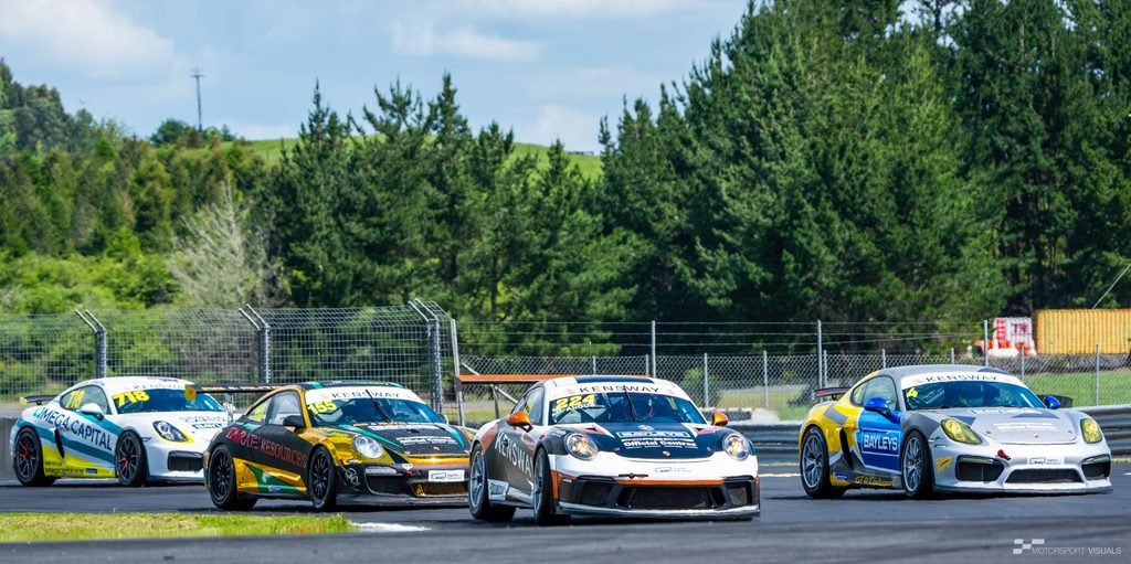 A dynamic action shot captures three race cars, specifically Porsche 911 models, lined up on a racetrack during an outdoor competition. The cars are branded with 'KENSWAY' and feature various liveries, suggesting a motorsport event. The background consists of dense green trees and a clear blue sky with scattered clouds, indicating pleasant weather conditions. The cars are poised on the asphalt track surface, ready for action.