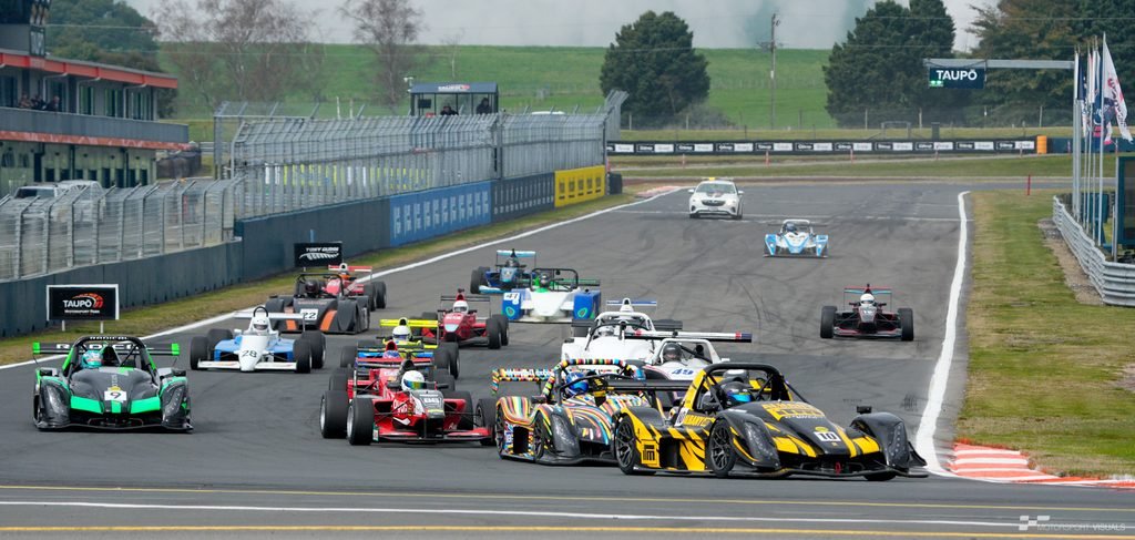 A group of race cars lined up on a racetrack during an overcast day, suggesting the start or practice session.