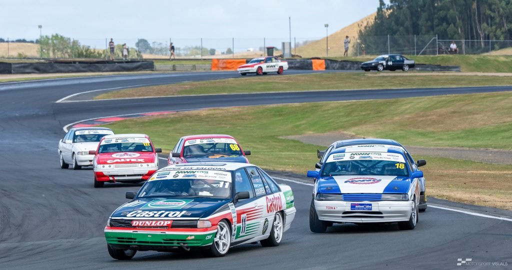 A race scene featuring several cars on a track under an overcast sky. The main focus is on three brightly liveried race cars in the foreground, with other vehicles and track infrastructure visible in the background.