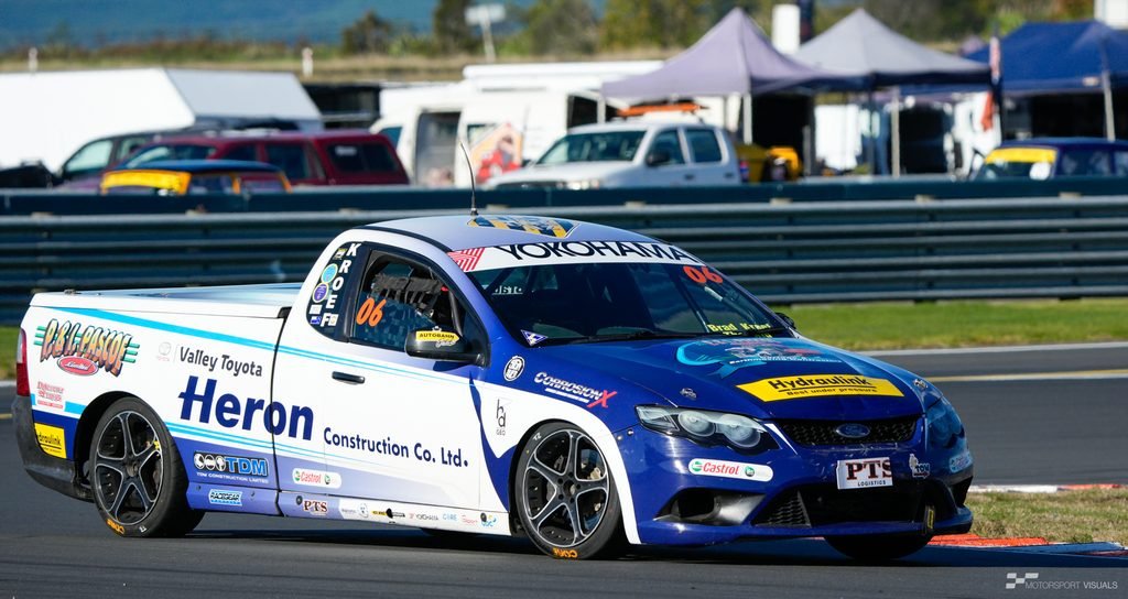 A low-angle action shot captures a race car, specifically a Ford Fiesta rally machine sponsored by various companies including Heron Construction Co. and Yokohama, speeding across an asphalt track. The car is painted in a vibrant blue livery with white accents and features racing decals, including the number 06. The background suggests a motorsports event setting with grandstands and tents under a bright, possibly overcast sky. The driver is visible inside the cockpit.
