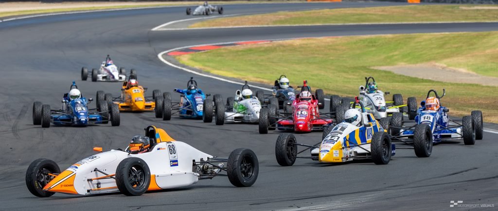 A group of race cars compete on a racetrack under clear skies. The lead white and orange racing car, number 88, is prominently featured in the foreground.