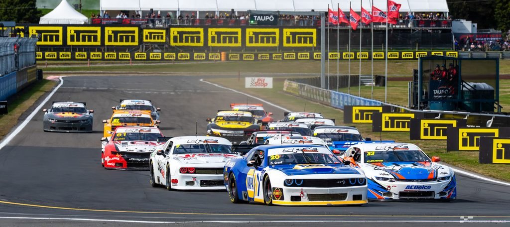 A group of race cars lined up on a track during a motorsports event, with grandstands and sponsor banners in the background.